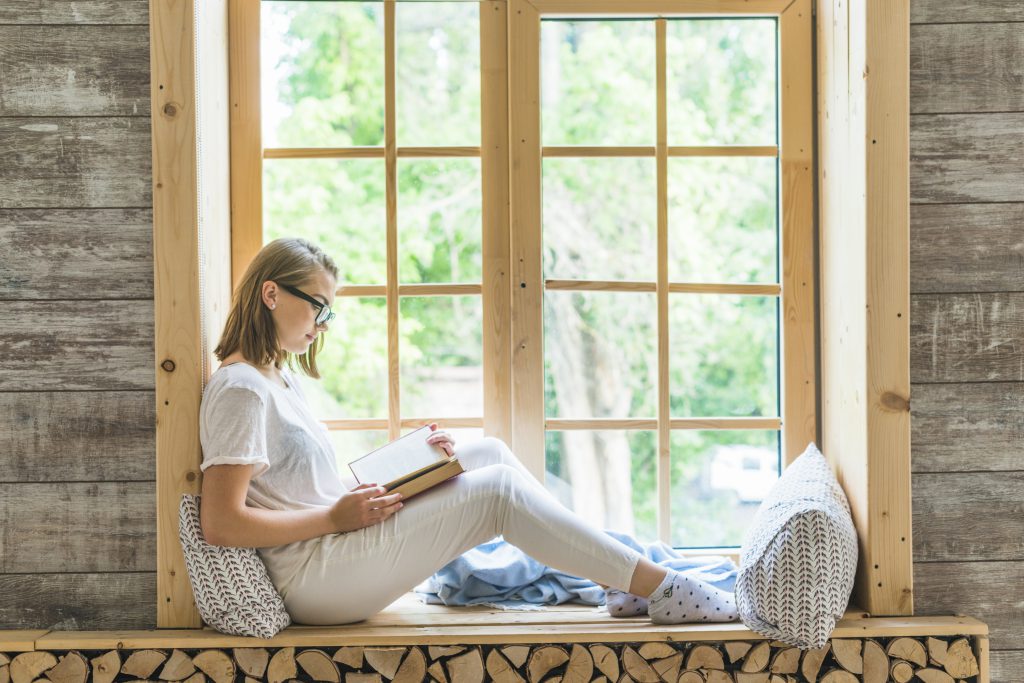 Kobieta siedząca przy dużym oknie drewnianym w domu – okna drewniane cena a komfort wnętrza.
