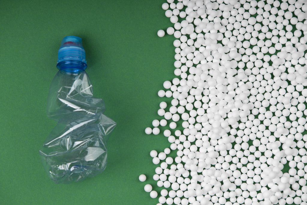 Granulat tworzywa PA11 poliamid obok zgniecionej plastikowej butelki na zielonym tle, symbolizujący recykling i zrównoważoną produkcję.