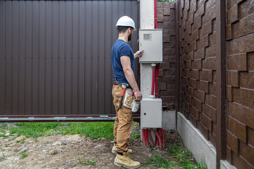Instalator w kasku i stroju roboczym konfiguruje napęd FAAC do bramy przesuwnej, otwierając skrzynkę sterowniczą.